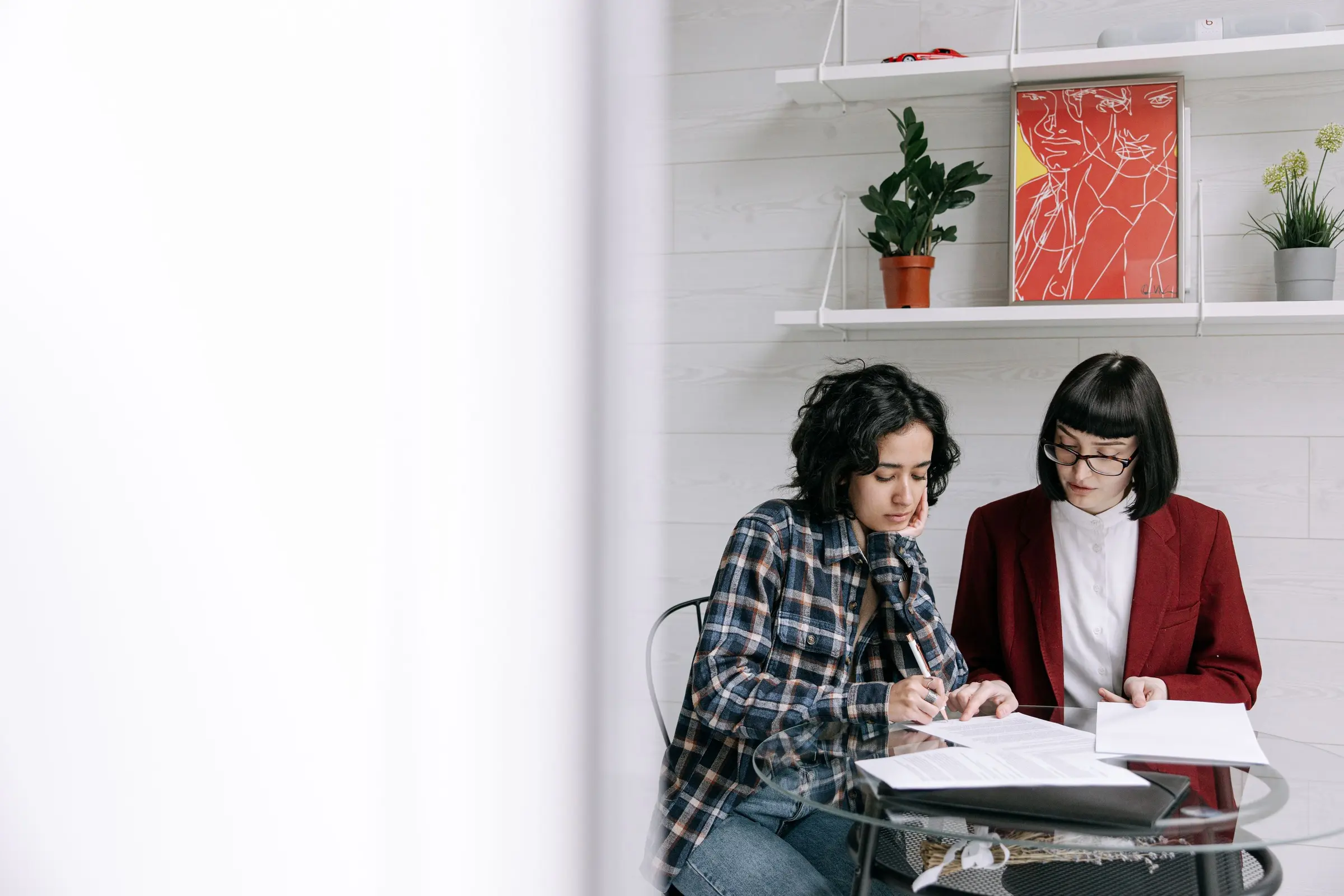 Illustrative photograph: two colleagues review documents at a glass table in daylight; one wears a red blazer